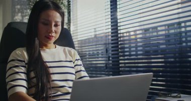 Bilgisayarında çalışan güzel Asyalı kadının kamera görüntüsü. Büyük panoramik pencerenin önünde panjurlarla oturan meşgul bir kız. Dijital cihaz kullanılıyor. Ekrana bakarken neşeyle gülümsüyor..