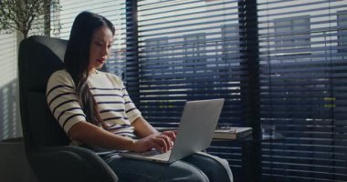 Ofis sandalyesinde otururken dizlerinin üstünde dizüstü bilgisayar tutan meşgul bir kadın. Metni klavyeye giriyorum. Meslektaşına mesaj yazıyor. Arka planda yarı kapalı panjurları olan büyük pencereler.