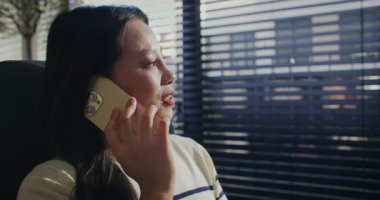 Asyalı bir kadının cep telefonuyla konuştuğu bir kamera görüntüsü. Panjurlarla panoramik pencereden bakıyorum. Uzak konuşmalar sırasında sevinçten sırıtarak. Çevrimiçi iletişim.