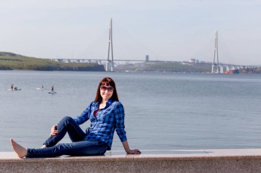 Deniz seti üzerinde rahat elbiseli hoş genç beyaz bir kadının portresi. Ünlü Russky Köprüsü arka plandadır. Rusya, Vladivostok, Russky Adası.