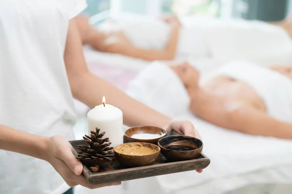 Massage therapist holding spa treatment set and hot scrub lotion with woman lying on spa bed prepared for spa massage in background. Luxury wellness, stress relief and rejuvenation concept.