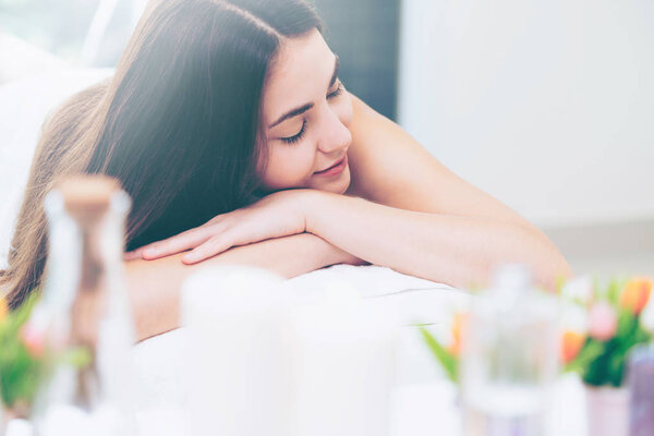 Relaxed woman lying on spa bed for aromatherapy massage in luxury spa with blurred foreground of spa treatment set including aromatic oil, candle and herbal scrub. Wellness and healing concept.