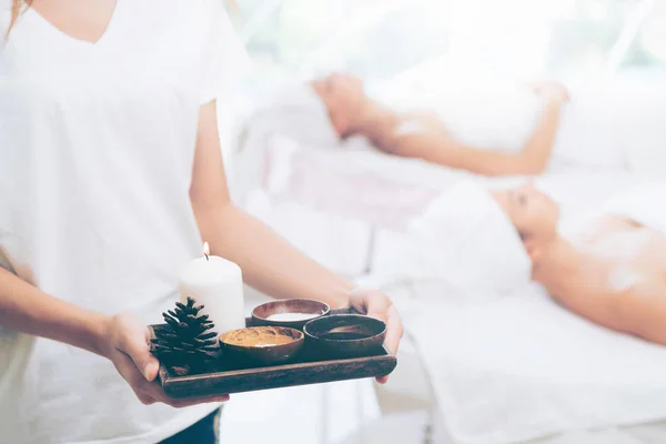 Massage therapist holding spa treatment set and hot scrub lotion with woman lying on spa bed prepared for spa massage in background. Luxury wellness, stress relief and rejuvenation concept.