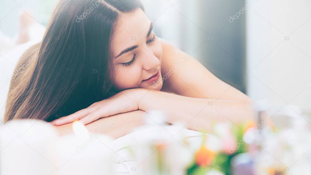 Relaxed woman lying on spa bed for aromatherapy massage in luxury spa with blurred foreground of spa treatment set including aromatic oil, candle and herbal scrub. Wellness and healing concept.