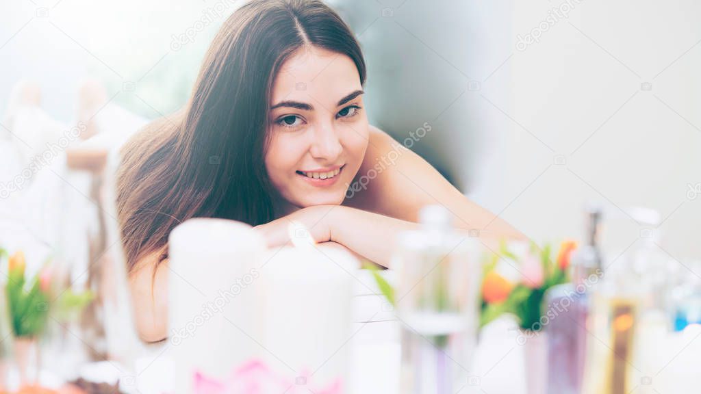 Relaxed woman lying on spa bed for aromatherapy massage in luxury spa with blurred foreground of spa treatment set including aromatic oil, candle and herbal scrub. Wellness and healing concept.