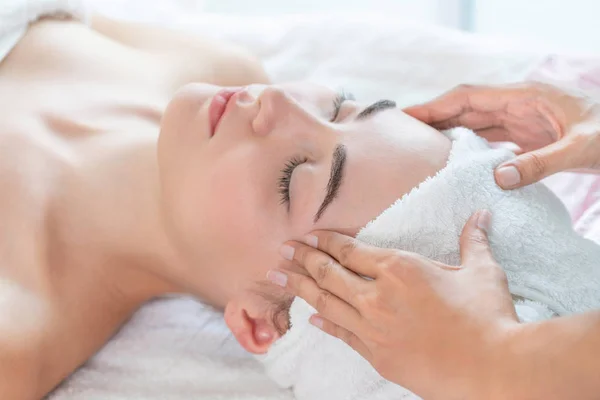 Relaxed woman lying on spa bed for facial and head massage spa treatment by massage therapist in a luxury spa resort. Wellness, stress relief and rejuvenation concept.