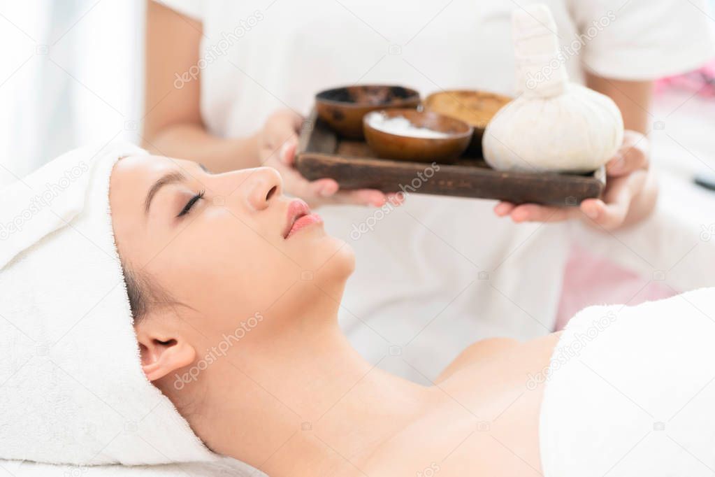 Relaxed woman lying on spa bed prepared for spa massage with therapist holding spa treatment set in background. Luxury wellness, stress relief and rejuvenation concept.