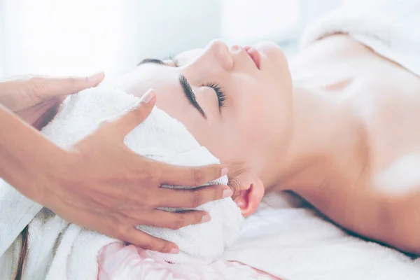 Relaxed woman lying on spa bed for facial and head massage spa treatment by massage therapist in a luxury spa resort. Wellness, stress relief and rejuvenation concept.