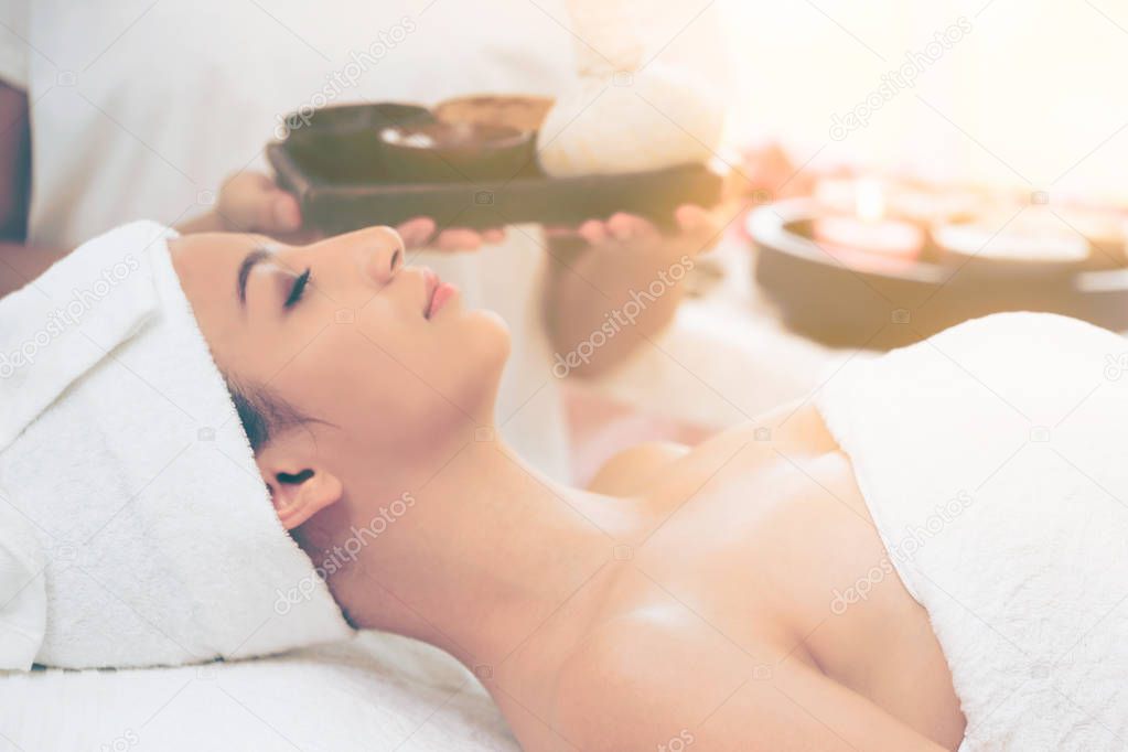 Relaxed woman lying on spa bed prepared for spa massage with therapist holding spa treatment set in background. Luxury wellness, stress relief and rejuvenation concept.