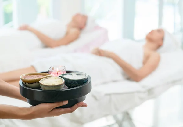 Massage therapist holding spa treatment set and hot scrub lotion with woman lying on spa bed prepared for spa massage in background. Luxury wellness, stress relief and rejuvenation concept.