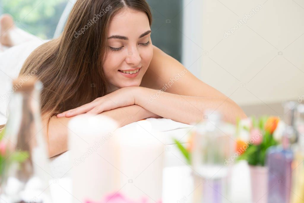 Relaxed woman lying on spa bed for aromatherapy massage in luxury spa with blurred foreground of spa treatment set including aromatic oil, candle and herbal scrub. Wellness and healing concept.