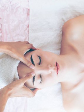 Lüks bir spa merkezinde masaj terapisti tarafından yüz masajı ve spa tedavisi için kaplıcaya uzanmış rahat bir kadın. Sağlık, stres atma ve gençleştirme kavramı.