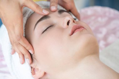 Lüks bir spa merkezinde masaj terapisti tarafından yüz masajı ve spa tedavisi için kaplıcaya uzanmış rahat bir kadın. Sağlık, stres atma ve gençleştirme kavramı.