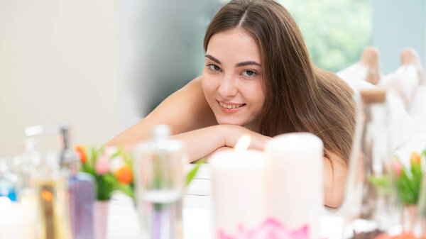 Relaxed woman lying on spa bed for aromatherapy massage in luxury spa with blurred foreground of spa treatment set including aromatic oil, candle and herbal scrub. Wellness and healing concept.