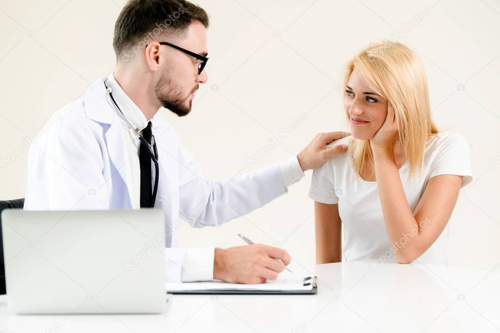 Male doctor talks to female patient in hospital office while writing on the patients health record on the table. Healthcare and medical service.