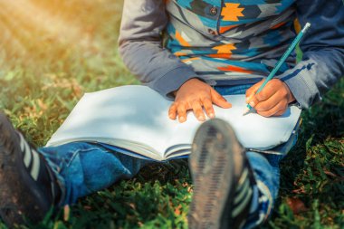 Küçük çocuk temel üstünde bir defter kalem ile parkta yeşil çimenlerin üzerinde otururken yazma. Çocuk eğitim kavramı.