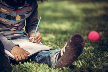 Küçük çocuk temel üstünde bir defter kalem ile parkta yeşil çimenlerin üzerinde otururken yazma. Çocuk eğitim kavramı.