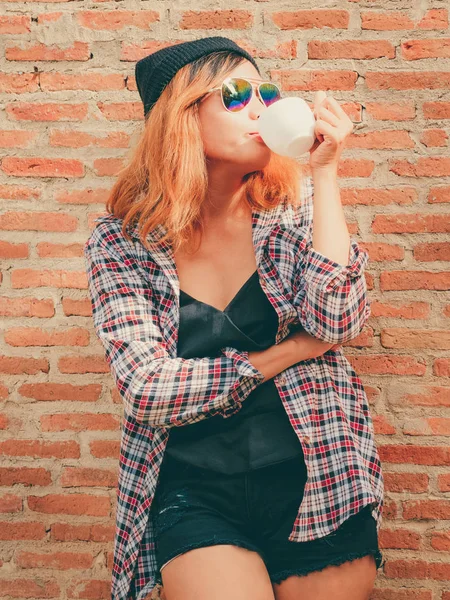 Onun elinde kahve fincanı ile şehirdeki sokak duvara karşı duran mutlu genç hipster kadın.