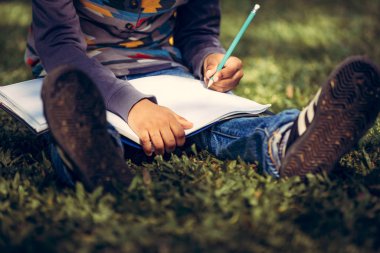 Küçük çocuk temel üstünde bir defter kalem ile parkta yeşil çimenlerin üzerinde otururken yazma. Çocuk eğitim kavramı.