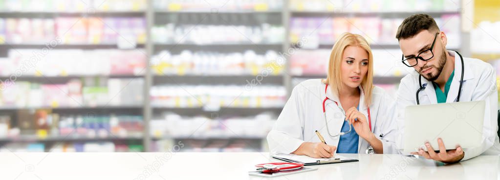 Pharmacist working with laptop computer at the office while having discussion with another pharmacist in the pharmacy. Medical healthcare and pharmaceutical service.