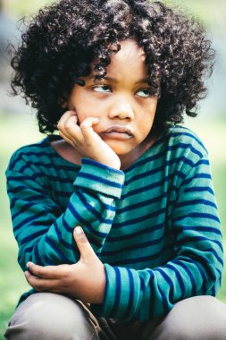 Unhappy bored little african american kid sitting in the park. The boy showing negative emotion. Child trouble concept. clipart