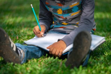 Küçük çocuk temel üstünde bir defter kalem ile parkta yeşil çimenlerin üzerinde otururken yazma. Çocuk eğitim kavramı.