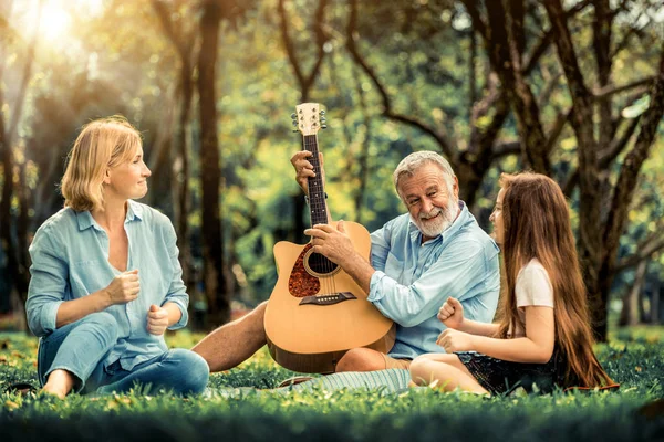 Happy family play guitar and sing together while sitting in the park in summer. Concept of family bonding by music.