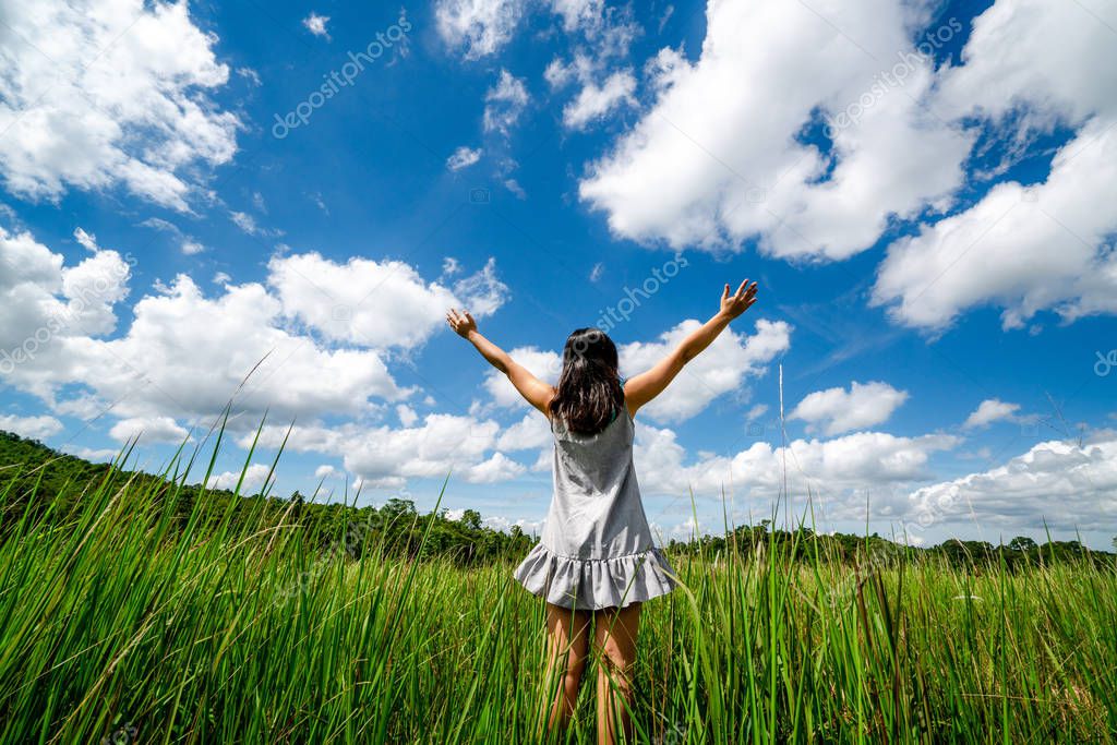 Young beautiful woman on green grass meadow spreading arms to the blue sunny sky on summer day. Getaway and meditation concept.