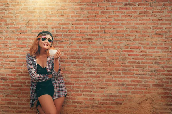 Onun elinde kahve fincanı ile şehirdeki sokak duvara karşı duran mutlu genç hipster kadın.