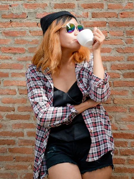 Onun elinde kahve fincanı ile şehirdeki sokak duvara karşı duran mutlu genç hipster kadın.