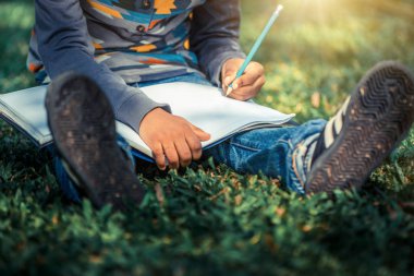 Küçük çocuk temel üstünde bir defter kalem ile parkta yeşil çimenlerin üzerinde otururken yazma. Çocuk eğitim kavramı.