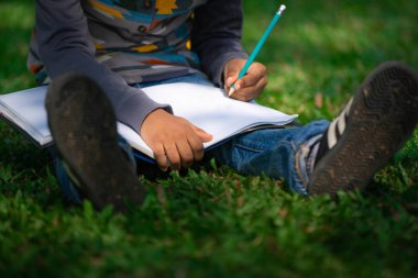 Küçük çocuk temel üstünde bir defter kalem ile parkta yeşil çimenlerin üzerinde otururken yazma. Çocuk eğitim kavramı.