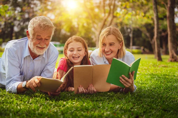 Happy family read books together and lying on green grass in public park. Little girl kid learning with mother and father in outdoors garden. Education and family lifestyle.