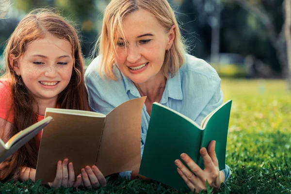 Happy family read books together and lying on green grass in public park. Little girl kid learning with mother in outdoors garden. Education and family lifestyle.