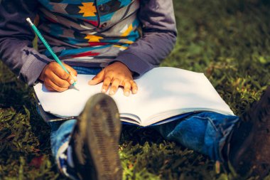 Küçük çocuk temel üstünde bir defter kalem ile parkta yeşil çimenlerin üzerinde otururken yazma. Çocuk eğitim kavramı.