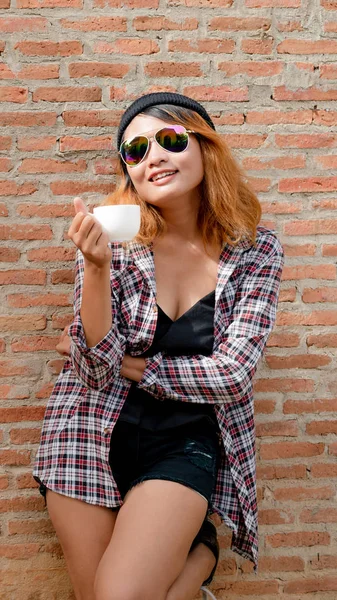 彼女の手でコーヒー カップを持つ町通りのレンガ壁に立っている幸せな若いヒップな女性 — ストック写真