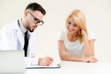Male doctor talks to female patient in hospital office while writing on the patients health record on the table. Healthcare and medical service. clipart
