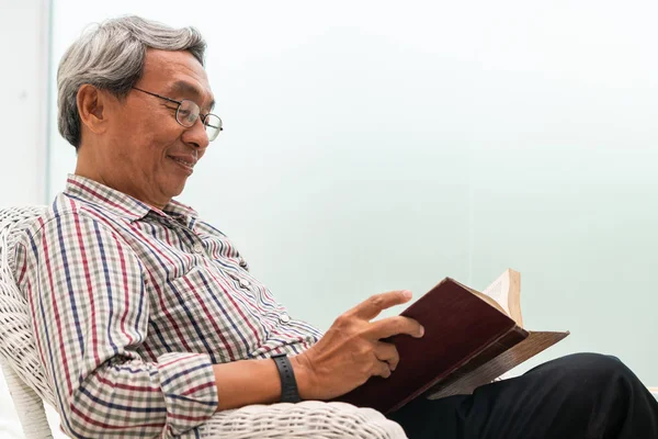 Zkušený asijský muž čte knížku na židli doma. — Stock fotografie