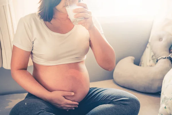 Happy pregnant woman drinking milk in glass at home while taking care of child. Young expecting mother holding baby in pregnant belly. Calcium food nutrition for strong bones during pregnancy.