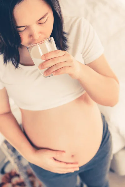Happy pregnant woman drinking milk in glass at home while taking care of child. Young expecting mother holding baby in pregnant belly. Calcium food nutrition for strong bones during pregnancy.