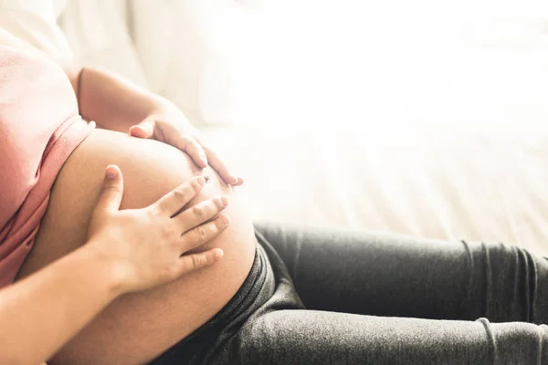 Pregnant woman feeling happy at home while taking care of child. Young expecting mother holding baby in pregnant belly. Maternity prenatal care and woman pregnancy concept.