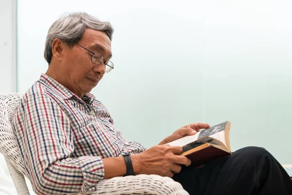 Senior Aziatische man lezen boek op stoel thuis. — Stockfoto