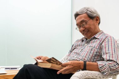 Mutlu üst düzey Asyalı adam evde oturma odasında sandalyede kitap okuyor. Emeklilik yaşam tarzı ve yaşlanma toplum kavramı.