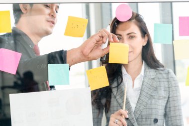 İş adamları ofisteki proje planlama kurulunda çalışıyor ve proje gelişimini analiz etmek için iş arkadaşlarıyla sohbet ediyorlar. Düzenli olması için cam duvara yapıştırılmış yapışkan notlar kullanıyorlar. .