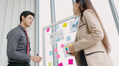 İş adamları ofisteki proje planlama kurulunda çalışıyor ve proje gelişimini analiz etmek için iş arkadaşlarıyla sohbet ediyorlar. Düzenli olması için cam duvara yapıştırılmış yapışkan notlar kullanıyorlar. .