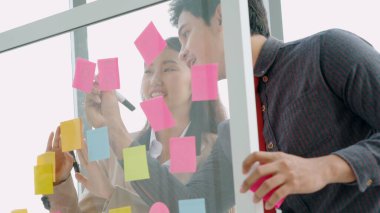 İş adamları ofisteki proje planlama kurulunda çalışıyor ve proje gelişimini analiz etmek için iş arkadaşlarıyla sohbet ediyorlar. Düzenli olması için cam duvara yapıştırılmış yapışkan notlar kullanıyorlar. .