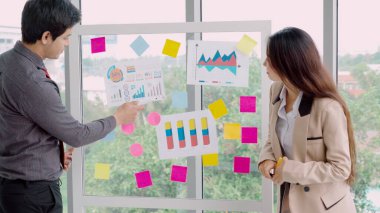 İş adamları ofisteki proje planlama kurulunda çalışıyor ve proje gelişimini analiz etmek için iş arkadaşlarıyla sohbet ediyorlar. Düzenli olması için cam duvara yapıştırılmış yapışkan notlar kullanıyorlar. .