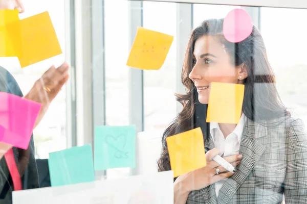 Business people work on project planning board in office and having conversation with coworker friend to analyze project development . They use sticky notes posted on glass wall to make it organized .