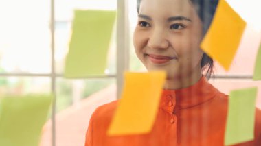 İş adamları ofisteki proje planlama kurulunda çalışıyor ve proje gelişimini analiz etmek için iş arkadaşlarıyla sohbet ediyorlar. Düzenli olması için cam duvara yapıştırılmış yapışkan notlar kullanıyorlar. .