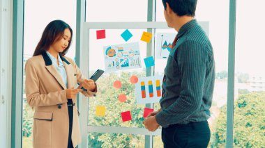 İş adamları ofisteki proje planlama kurulunda çalışıyor ve proje gelişimini analiz etmek için iş arkadaşlarıyla sohbet ediyorlar. Düzenli olması için cam duvara yapıştırılmış yapışkan notlar kullanıyorlar. .